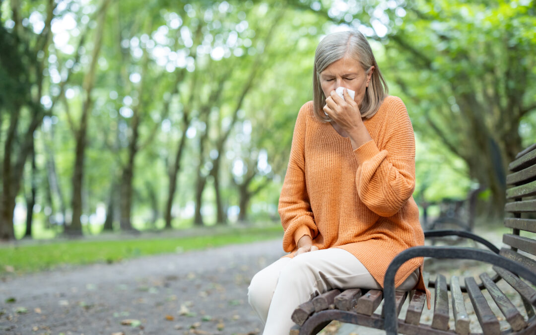 Senior woman in orange sweater sitting on park bench sneezing into tissue. Outdoor setting during autumn signifies allergy or cold symptoms, highlighting seasonal health issues and colder weather.